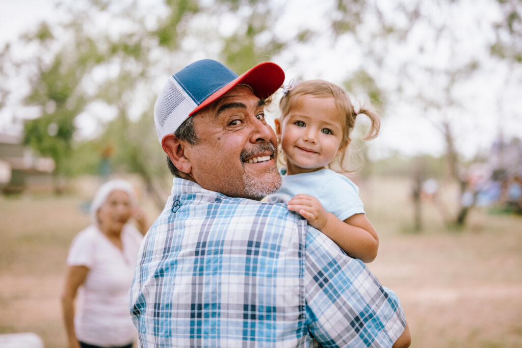 A grandfather in a trucker cap holding his young granddaughter on his shoulder, both smiling