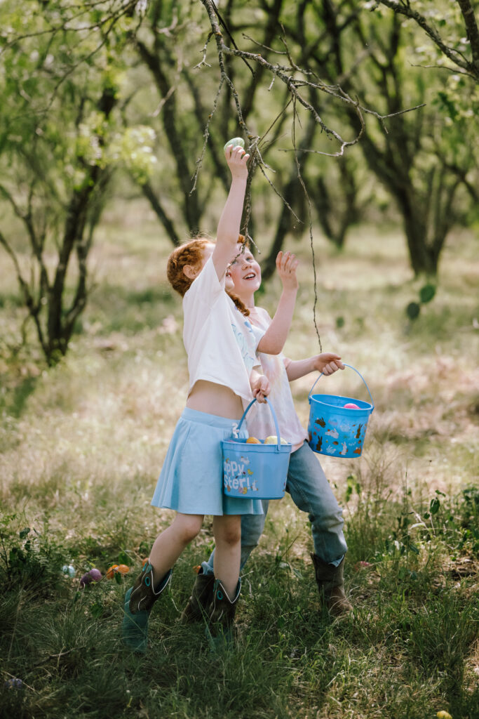 Two children in cowboy boots reaching up into a tree with light blue Easter buckets at golden hour